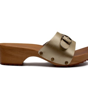 May include: A pair of gold leather heeled clogs with a buckle closure. The clogs have a wooden platform and a textured sole.