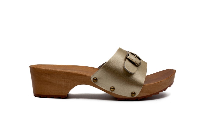 May include: A pair of gold leather heeled clogs with a buckle closure. The clogs have a wooden platform and a textured sole.