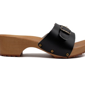 Puede incluir: Zueco de tacón de cuero negro con hebilla y plataforma de madera.