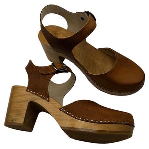 Woman wearing handmade brown leather wooden clogs with ankle strap and block wood heel, styled with cream linen dress against warm neutral background.