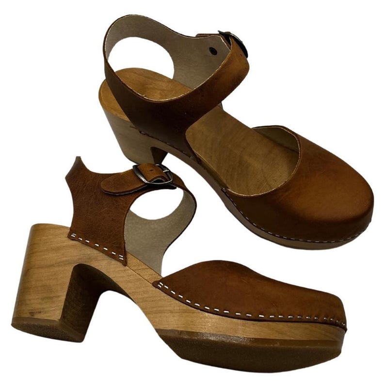 Woman wearing handmade brown leather wooden clogs with ankle strap and block wood heel, styled with cream linen dress against warm neutral background.
