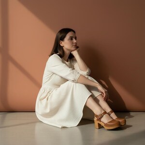 Woman wearing handmade brown leather wooden clogs with ankle strap and block wood heel, styled with cream linen dress against warm neutral background.