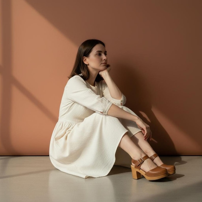 Woman wearing handmade brown leather wooden clogs with ankle strap and block wood heel, styled with cream linen dress against warm neutral background.