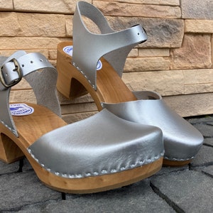 May include: A pair of silver leather clogs with wooden heels and a strap around the ankle. The clogs have a stitched edge and a light wood sole.