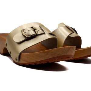 May include: A pair of gold leather heeled clogs with a buckle closure. The clogs have a wooden base and a textured sole.