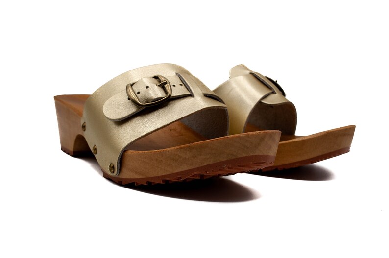 May include: A pair of gold leather heeled clogs with a buckle closure. The clogs have a wooden base and a textured sole.