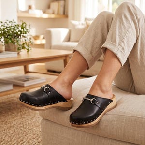 A person sits on a light-colored sofa, wearing beige pants and black leather clogs with silver-toned metal buckles and wooden soles.