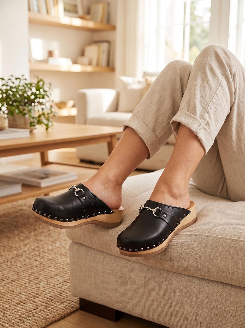 A person sits on a light-colored sofa, wearing beige pants and black leather clogs with silver-toned metal buckles and wooden soles.