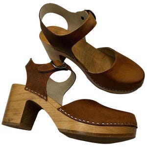 Woman wearing handmade brown leather wooden clogs with ankle strap and block wood heel, styled with cream linen dress against warm neutral background.