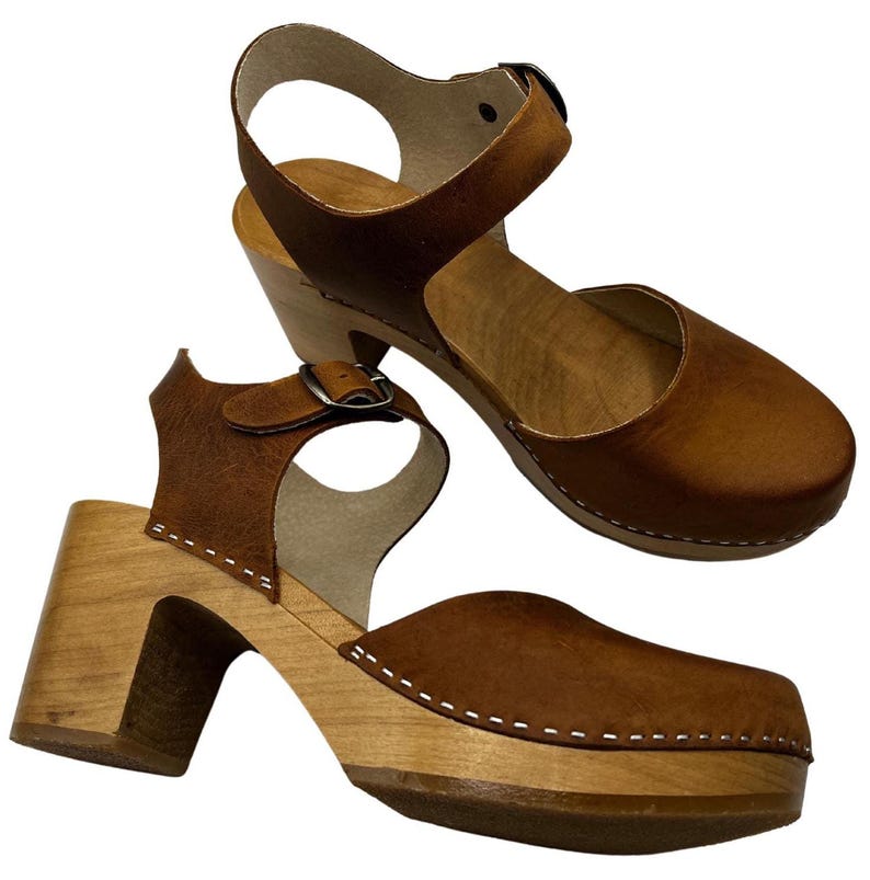 Woman wearing handmade brown leather wooden clogs with ankle strap and block wood heel, styled with cream linen dress against warm neutral background.