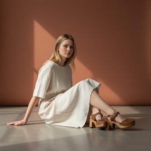 Woman wearing handmade brown leather wooden clogs with ankle strap and block wood heel, styled with cream linen dress against warm neutral background.