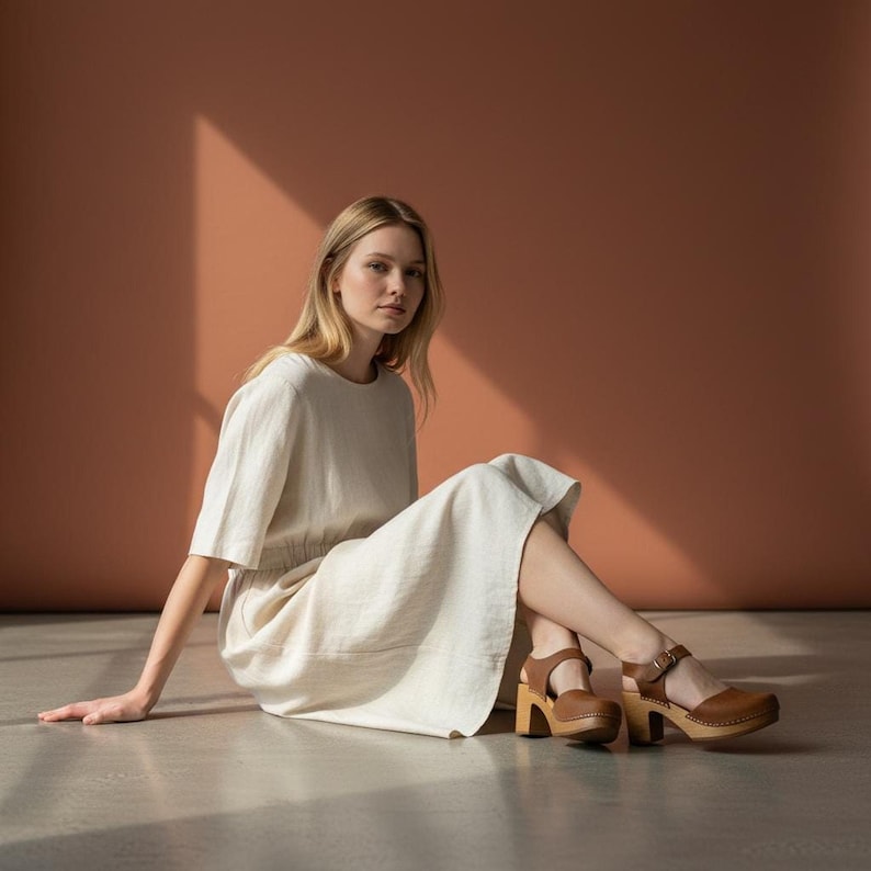Woman wearing handmade brown leather wooden clogs with ankle strap and block wood heel, styled with cream linen dress against warm neutral background.