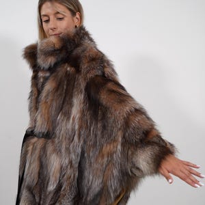 May include: A luxurious fur cape in shades of brown, silver, and gold. The cape features a high collar and a flowing, open design. The fur appears soft and plush, creating a glamorous and elegant look. The garment is worn over a black outfit.