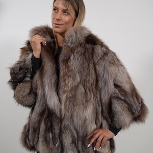 Könnte beinhalten: Ein luxuriöser, braun-silberner Pelzmantel mit breitem Kragen und kurzen Ärmeln. Der Mantel hat ein weiches, strukturiertes Aussehen und wird über einem schwarzen Oberteil und Leggings getragen. Das Fell scheint von einem Fuchs oder einer ähnlichen Art zu stammen.