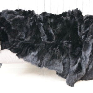 May include: A black faux fur throw blanket draped over a light grey sofa. The blanket is made of soft, fluffy fur and has a luxurious feel.