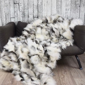 Può includere: Una coperta in pelliccia sintetica bianca e grigia drappeggiata su un divano grigio. La coperta è realizzata in pelliccia morbida e soffice e ha una sensazione lussuosa.