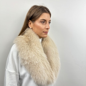 May include: A white fur collar with a soft, fluffy texture. The collar is worn around the neck of a person wearing a white sweater.