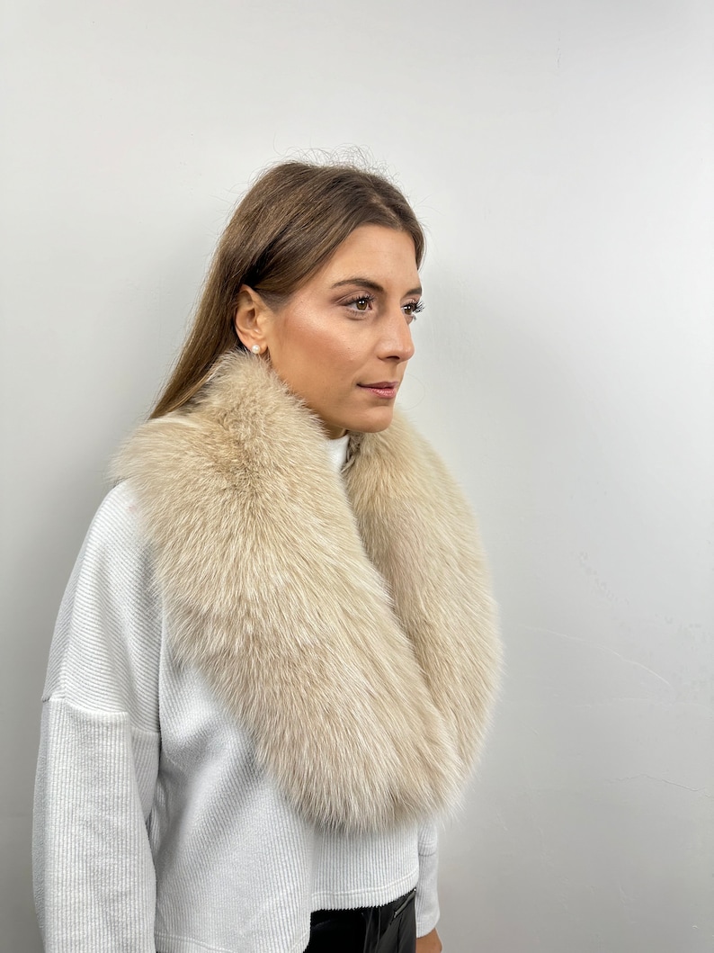 May include: A white fur collar with a soft, fluffy texture. The collar is worn around the neck of a person wearing a white sweater.