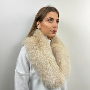 May include: A white fur collar with a soft, fluffy texture. The collar is worn around the neck of a person wearing a white sweater.