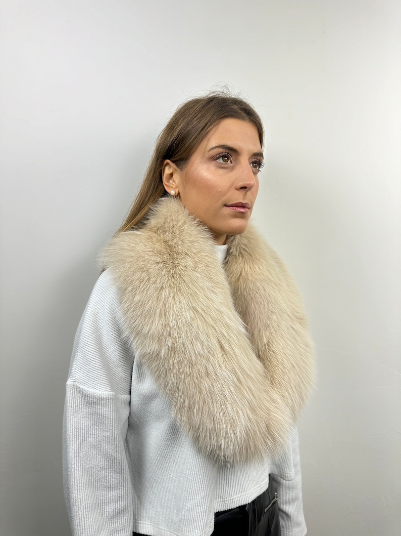 May include: A white fur collar with a soft, fluffy texture. The collar is worn around the neck of a person wearing a white sweater.