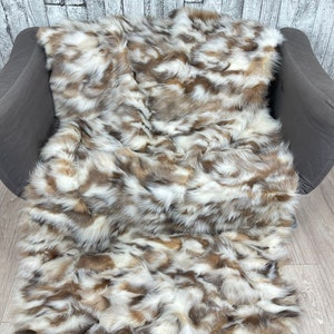 May include: A soft, fluffy throw blanket made from fox fur. The fur is a mix of white, brown, and black, creating a unique and luxurious look. The blanket is draped over a gray couch.