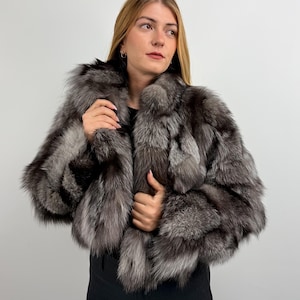 Puede incluir: Una chaqueta corta de piel de zorro plateado. La chaqueta presenta una mezcla de piel plateada y gris oscuro, creando una apariencia texturizada. La modelo lleva un top negro y pantalones negros. El fondo es una pared blanca lisa.