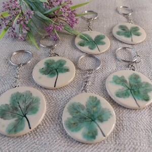 Può includere: Un set di sette portachiavi con un design a foglia verde pressata su uno sfondo in ceramica bianca. Ogni portachiavi ha un anello e una catena in metallo argentato.