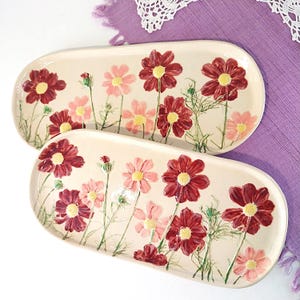 Könnte beinhalten: Zwei ovale Servierplatten aus Keramik mit floralem Design. Die Platten sind mit rosa und roten Kosmeen auf cremefarbenem Hintergrund verziert.