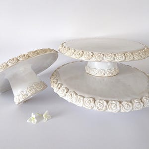 Könnte beinhalten: Drei weiße Keramik-Tortenplatten mit einem erhabenen Rosenmuster an den Rändern. Die Platten sind unterschiedlich groß und werden übereinander gestapelt.