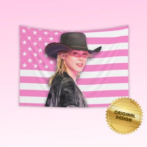 May include: A rectangular wall tapestry featuring a portrait of a person with blonde hair wearing a dark cowboy hat and a dark jacket. The background is a flag pattern with horizontal pink and white stripes and white stars. A gold seal reads ORIGINAL DESIGN.
