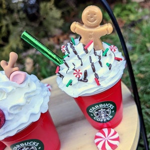 May include: Two red cups with white whipped cream and chocolate drizzle topped with candy canes and sprinkles. One cup has a gingerbread man on top and the other has a reindeer. Both cups have the Starbucks logo and the text "Coffee".