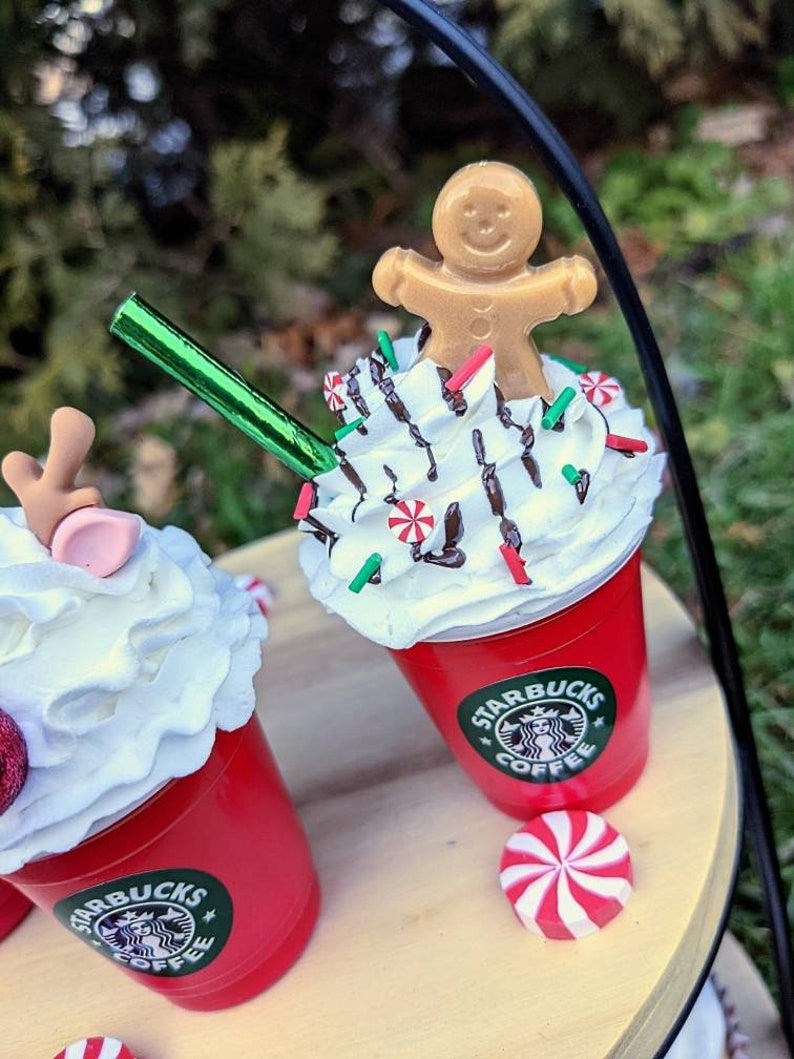 May include: Two red cups with white whipped cream and chocolate drizzle topped with candy canes and sprinkles. One cup has a gingerbread man on top and the other has a reindeer. Both cups have the Starbucks logo and the text "Coffee".