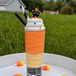 Puede incluir: Un vaso alto y transparente lleno de capas de crema texturizada negra, amarilla y naranja, cubierto con nata montada blanca, chispas de colores, dos pajitas a rayas blancas y negras y una pequeña figura de gato negro con un sombrero de calabaza.