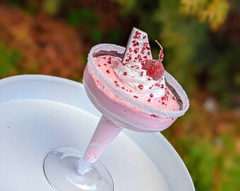 Puede incluir: Un postre rosa con crema batida blanca y chispas rojas en una copa de vidrio transparente sobre un plato blanco. El postre está cubierto con un trozo de chocolate y una cereza roja.
