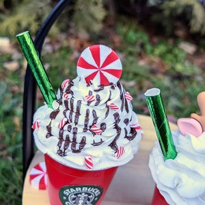 May include: Two miniature red cups with white whipped cream, chocolate drizzle, and candy canes. The cup on the left has a red and white peppermint candy on top and a green straw. The cup on the right has a green straw and a reindeer head on top. Both cups have the Starbucks logo on the side.