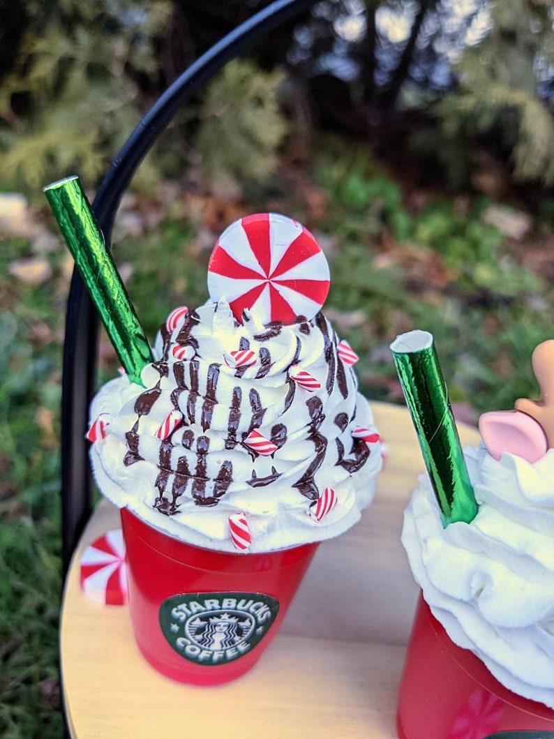 May include: Two miniature red cups with white whipped cream, chocolate drizzle, and candy canes. The cup on the left has a red and white peppermint candy on top and a green straw. The cup on the right has a green straw and a reindeer head on top. Both cups have the Starbucks logo on the side.