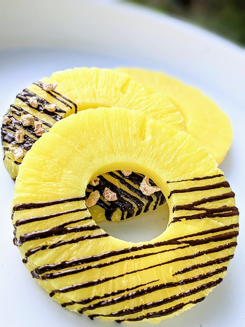 May include: Two pineapple rings topped with chocolate drizzle and chopped nuts. The pineapple rings are arranged on a white plate.