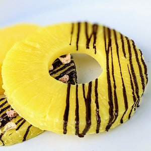 May include: Two pineapple rings with chocolate drizzled on top. The pineapple rings are on a white surface.