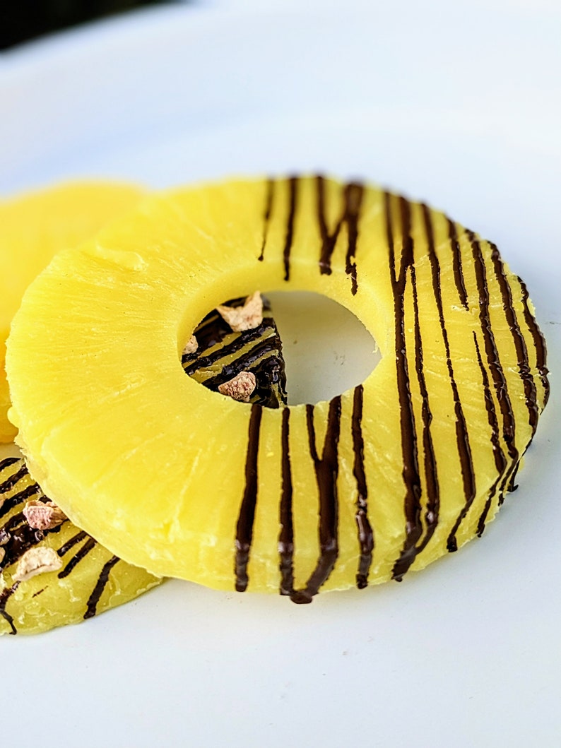 May include: Two pineapple rings with chocolate drizzled on top. The pineapple rings are on a white surface.