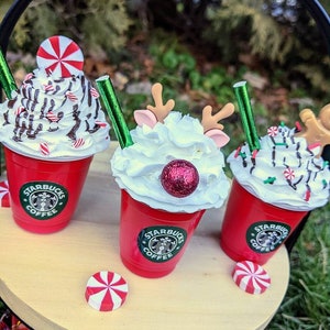 May include: Three red Starbucks cups with whipped cream and holiday themed toppings. The cups are on a wooden tiered tray. The toppings include a candy cane, a red and white peppermint, a red glitter ball, and a reindeer head.