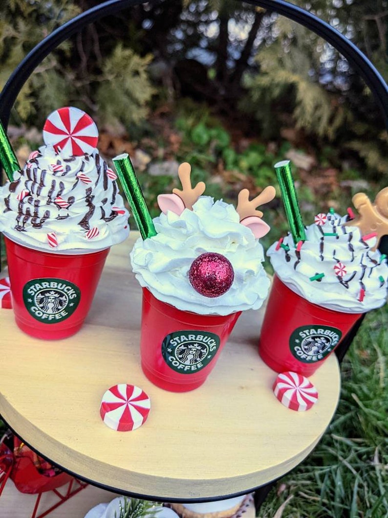 May include: Three red Starbucks cups with whipped cream and holiday themed toppings. The cups are on a wooden tiered tray. The toppings include a candy cane, a red and white peppermint, a red glitter ball, and a reindeer head.