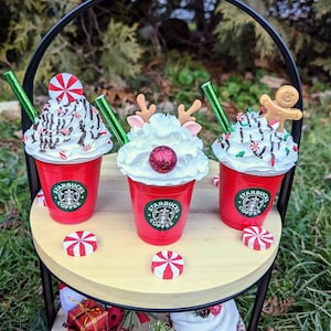 May include: Three festive Starbucks coffee cups with whipped cream and holiday decorations, including a reindeer, a gingerbread man, and a peppermint swirl. The cups are displayed on a two-tiered wooden stand with red and white peppermint candies.