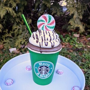Op de afbeelding: Een groene beker met het Starbucks-logo en de tekst "Merry & Bright" is bedekt met slagroom, chocoladesaus en een pepermuntje. Een groen rietje is in de beker gestoken. Verschillende pepermuntjes zijn rond de basis verspreid.