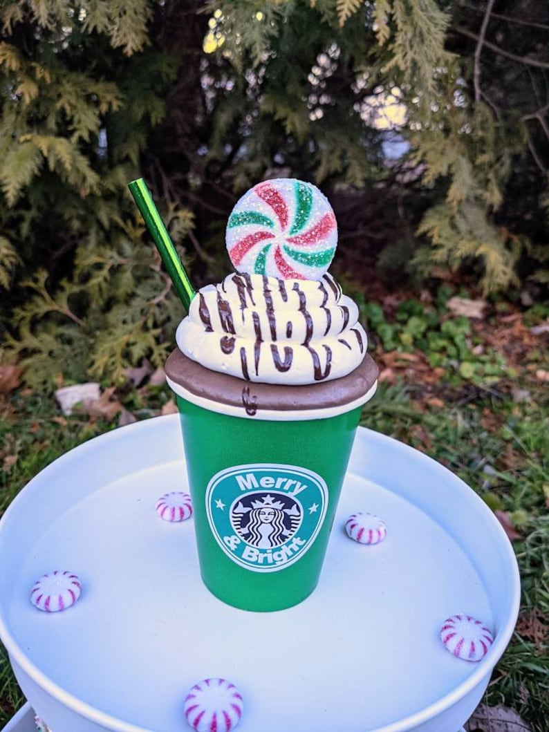 Op de afbeelding: Een groene beker met het Starbucks-logo en de tekst "Merry & Bright" is bedekt met slagroom, chocoladesaus en een pepermuntje. Een groen rietje is in de beker gestoken. Verschillende pepermuntjes zijn rond de basis verspreid.