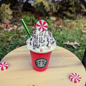 May include: A red Starbucks cup with a white whipped cream topping, chocolate drizzle, and candy cane sprinkles. A candy cane sits on top of the whipped cream. A green straw is in the cup. The cup is sitting on a wooden surface with three candy canes.