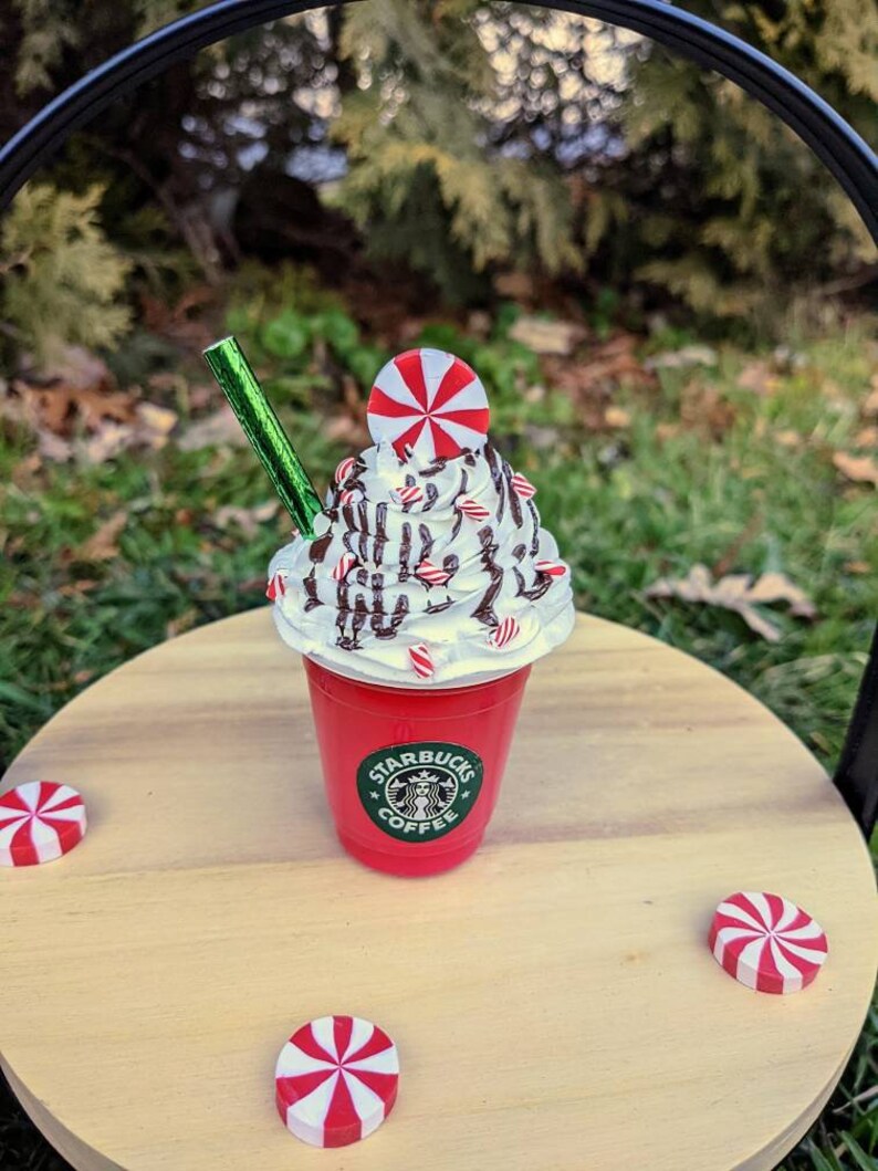 May include: A red Starbucks cup with a white whipped cream topping, chocolate drizzle, and candy cane sprinkles. A candy cane sits on top of the whipped cream. A green straw is in the cup. The cup is sitting on a wooden surface with three candy canes.