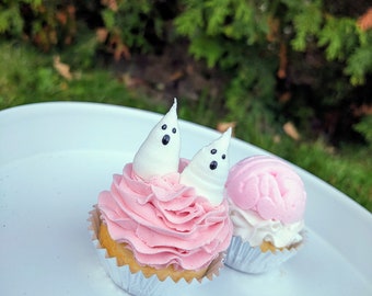 Könnte beinhalten: Zwei Cupcakes auf einem weißen Teller. Einer hat rosa Zuckerguss mit zwei weißen Geisterdekorationen mit schwarzen Punktaugen. Der andere hat rosa und weißen Zuckerguss. Beide sind in silbernen Folienförmchen.