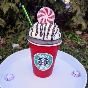 Op de afbeelding: Een feestelijke rode beker met een groen rietje, gegarneerd met slagroom, chocoladesaus en een pepermunt snoepje. De beker heeft een logo en de tekst "Merry & Bright". Verschillende pepermunt snoepjes zijn rond de basis geplaatst.