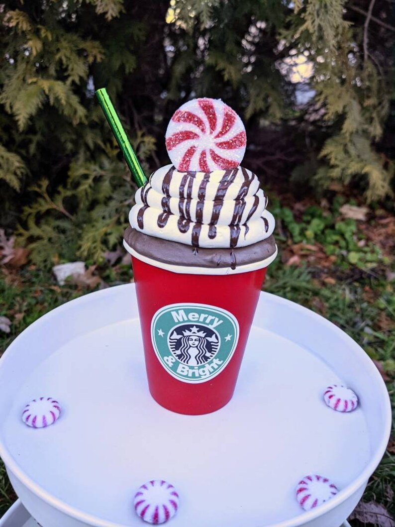 Op de afbeelding: Een feestelijke rode beker met een groen rietje, gegarneerd met slagroom, chocoladesaus en een pepermunt snoepje. De beker heeft een logo en de tekst "Merry & Bright". Verschillende pepermunt snoepjes zijn rond de basis geplaatst.