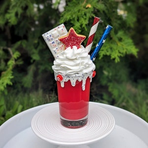 May include: A patriotic dessert in a clear glass, featuring whipped cream, a star-shaped biscuit, a white chocolate bar, and two straws. The glass is on a white cake stand. The dessert is red, white, and blue.